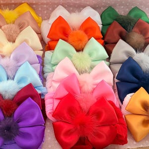Könnte beinhalten: Eine Sammlung von Satin-Haarschleifen in verschiedenen Farben, darunter Gelb, Orange, Grün, Rot, Lila, Blau und Pink. Jede Schleife hat in der Mitte einen flauschigen Pompon. Die Schleifen sind in einem Raster in einer hellrosa Schachtel angeordnet.