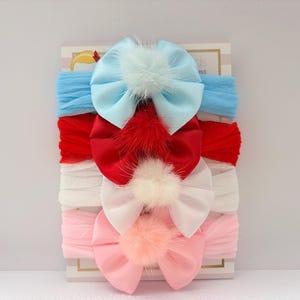 May include: A pack of four baby headbands in blue, red, white, and pink. Each headband has a satin bow with a fluffy pom-pom centre. The headbands are presented on a white and gold backing.