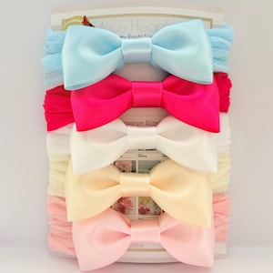 Puede incluir: Un conjunto de cinco diademas en una tarjeta de presentación, cada una con un lazo de satén liso. Los colores incluyen azul claro, rosa intenso, blanco, crema y rosa pálido. Cada lazo está unido a una diadema de tela fruncida a juego. Alice Headbands.