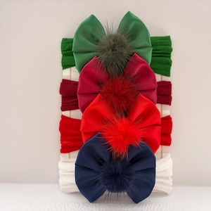 May include: A set of four headbands featuring large fabric bows in dark green, burgundy, bright red, and navy blue. Each bow is centred with a matching fluffy pom-pom. The headbands are displayed vertically on a white rectangular card backing.