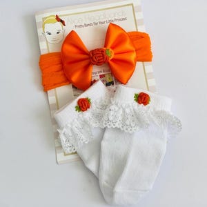 Puede incluir: Una diadema de nailon naranja con un gran lazo de satén y un pequeño adorno de calabaza bordada, junto con calcetines tobilleros blancos de canalé con ribete de encaje blanco y una pequeña calabaza naranja bordada en el puño.