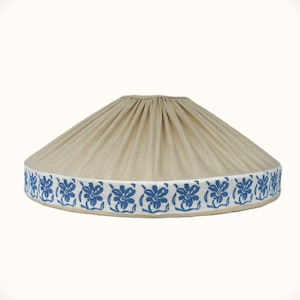 May include: A conical lampshade featuring a beige fabric body with vertical pleats. The bottom edge is finished with a white trim band decorated with a repeating pattern of blue floral motifs arranged in a continuous horizontal sequence.