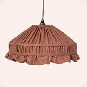 May include: A dusty rose pendant lamp shade featuring a pleated fabric design with a horizontal band detail and a ruffled bottom edge. The shade is suspended from a central cord with a metallic fixture at the top, showcasing a soft texture.
