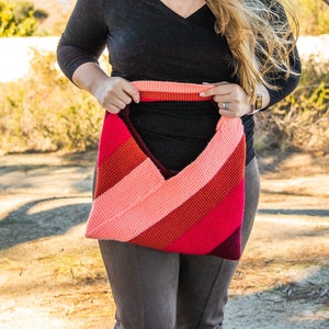 May include: A woman is holding a crocheted bag with a pink, red and burgundy striped design. The bag has a wide, open top and a slouchy shape.