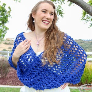 May include: A vibrant royal blue crochet shrug featuring an openwork, diamond-patterned stitch design. The garment is worn over a white top and includes a silver-toned necklace with a pink pendant and silver drop earrings as accessories.