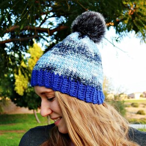 May include: A blue and white crocheted beanie with a faux fur pom pom. The beanie has a blue band at the bottom.
