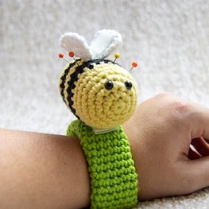 May include: A crocheted bee pincushion with black and yellow stripes, white wings, and black eyes. The bee is attached to a green crocheted wristband.