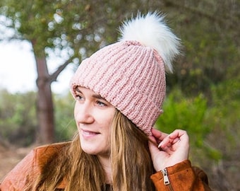 May include: A pink knitted beanie with a white pom pom. The beanie is worn by a person with long brown hair and a brown leather jacket.