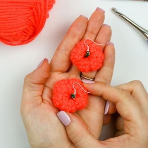 May include: A pair of crocheted earrings in a bright orange colour. The earrings are being held in a person's hands. The yarn used to make the earrings is in the background.