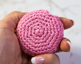 May include: A close-up of a pink crocheted circle, held in a hand with white nail polish. The circle is made of tightly woven yarn, creating a textured surface.