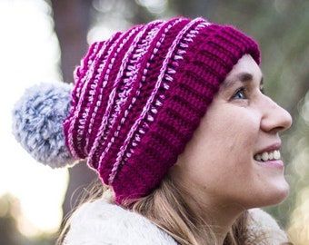 May include: A woman wearing a pink and white striped crochet beanie with a fluffy gray pom pom. The beanie has a contrasting white crocheted border. She is wearing a white faux fur jacket and looking up.