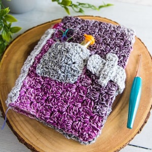May include: A crocheted purple and gray pouch with a pocket and a crocheted strap. The pouch is sitting on a wooden surface.