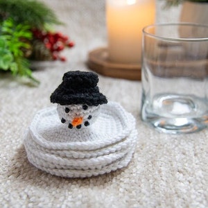 May include: A crocheted snowman coaster set with a black hat and orange nose. The coaster set is made of white yarn and is stacked on top of each other.