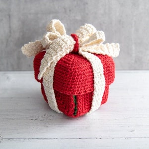 May include: A crocheted red gift box with a white crocheted bow. The box is sitting on a white wooden surface.