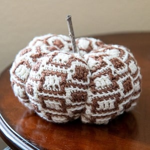 May include: A crocheted pumpkin with a brown and white geometric pattern. The pumpkin has a twig stem.
