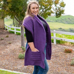 May include: A purple crochet cardigan with a shawl collar and a pocket on the front. The cardigan is worn over a black shirt and blue jeans.