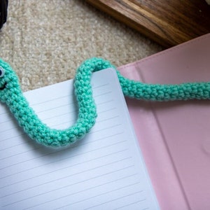 May include: A green crocheted snake bookmark with googly eyes. The bookmark is on a pink notebook.