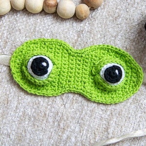 Puede incluir: Máscara para ojos de crochet verde con ojos blancos y negros. La máscara está unida a una cinta blanca.