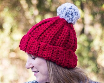 May include: A red crocheted beanie with a grey pom pom. The beanie is being worn by a person with long brown hair. The background is blurred and out of focus.