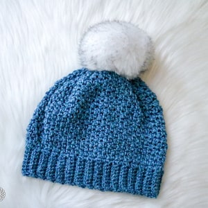Pode incluir: Um gorro de crochê azul com um pompom de pele sintética branca. O gorro é feito com um ponto texturizado.