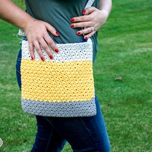 May include: A crocheted crossbody bag with a yellow, white, and gray striped design. The bag is worn by a person wearing blue jeans.