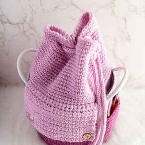 Puede incluir: Una bolsa de cord&oacute;n de crochet rosa con botones dorados. La bolsa est&aacute; sobre una superficie blanca.