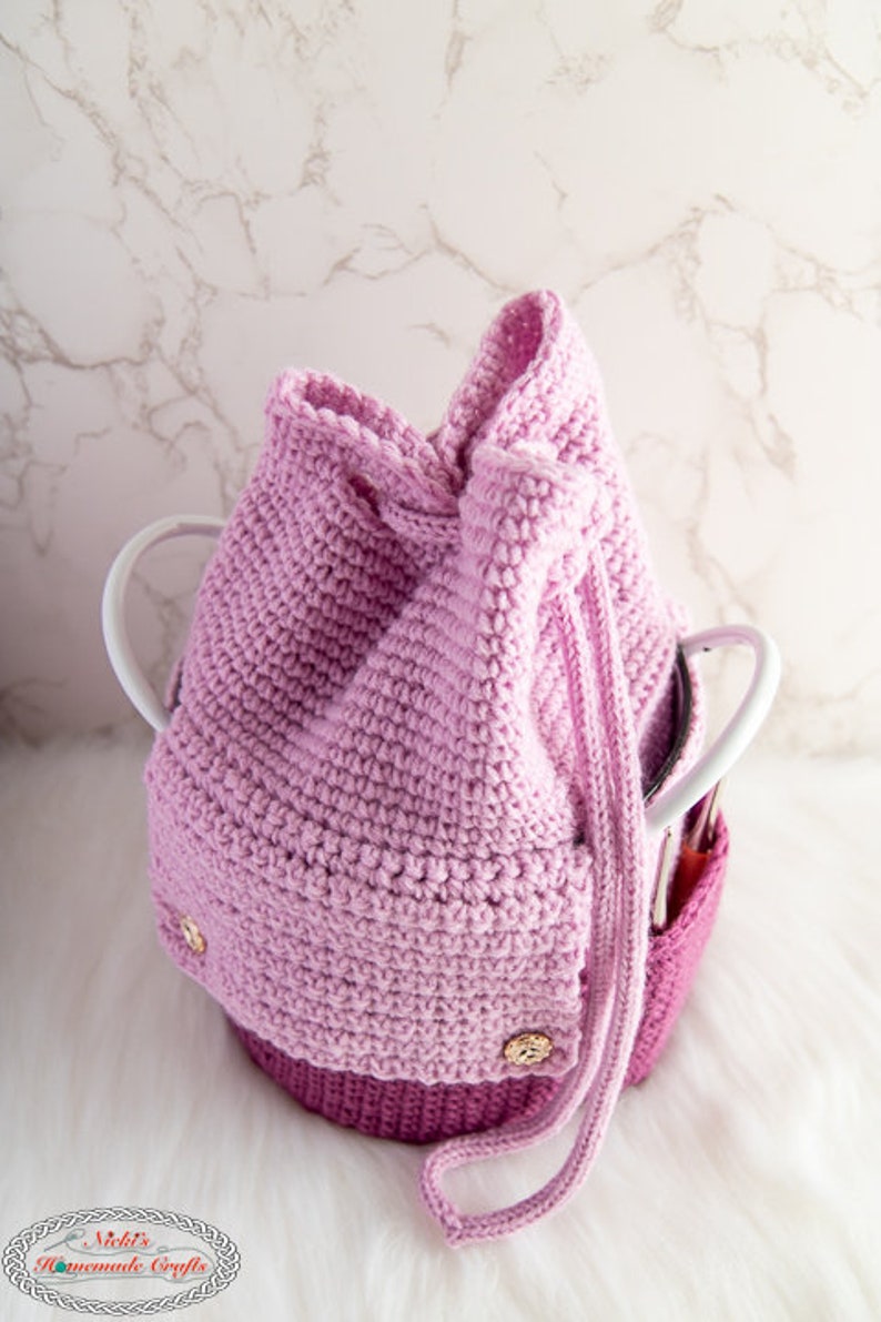 Puede incluir: Una bolsa de cord&oacute;n de crochet rosa con botones dorados. La bolsa est&aacute; sobre una superficie blanca.
