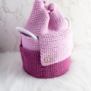 Puede incluir: Una bolsa de cord&oacute;n de ganchillo de color rosa y morado. La bolsa tiene un asa blanca y un bot&oacute;n dorado.