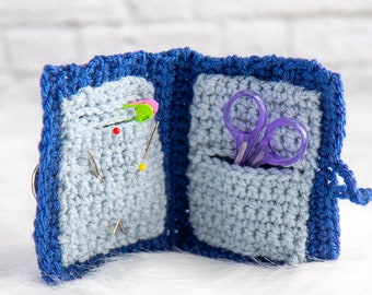 May include: A blue crocheted sewing needle and scissor holder with two compartments. One compartment has a variety of sewing needles and the other compartment has a pair of purple scissors.