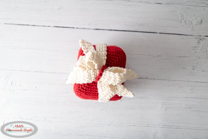 May include: A red crocheted gift box with a white crocheted bow on top. The box is sitting on a white wooden surface.