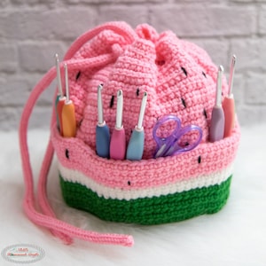 May include: A pink crocheted watermelon-shaped drawstring bag with a green and white stripe. The bag is filled with crochet hooks and a pair of purple scissors.
