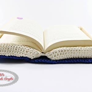 Pode incluir: Uma capa de livro de croch&ecirc; com um design branco, azul e marrom. A capa est&aacute; aberta para revelar um livro com p&aacute;ginas.