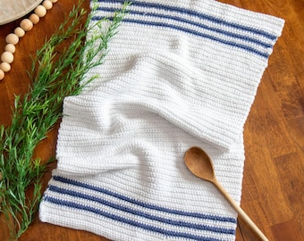 May include: White crocheted kitchen towel with blue stripes. The towel is folded and lying on a wooden table with a wooden spoon.