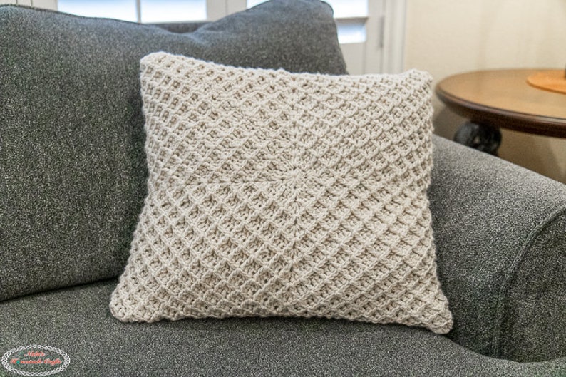 Puede incluir: Un coj&iacute;n de punto de ganchillo beige con un patr&oacute;n de diamantes sobre un sof&aacute; gris.