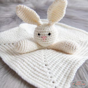 May include: A handmade, off-white crochet bunny security blanket. The bunny has long ears, a pink nose, and black eyes. The blanket is square with a textured stitch pattern, perfect for a baby or toddler.