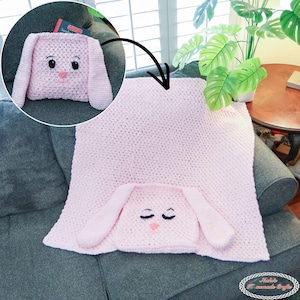May include: A handmade pink crochet bunny blanket and pillow set. The blanket features a hood with bunny ears and closed eyes. The pillow, in a circular inset, has a bunny face with black eyes and a pink nose. The set is displayed on a gray couch.