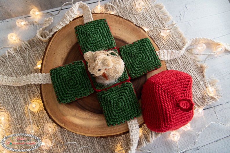 May include: A crocheted green gift box with a crocheted red ornament. The gift box is open and a small crocheted animal is inside. The box and ornament are on a wooden surface with a burlap background.