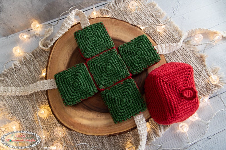 May include: A crocheted Christmas gift box set with a green box and a red ornament. The box is made of four squares and the ornament is round. The set is on a wooden surface with a white background.
