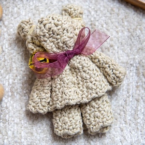 May include: A handmade, beige crochet teddy bear with a purple ribbon and a small gold bell. The bear is wearing a dress and has a textured appearance. The image is of a craft item.
