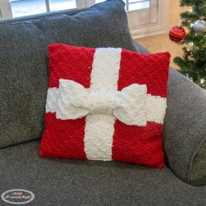 May include: A red and white crocheted cushion shaped like a wrapped present with a large white bow. The cushion is sitting on a grey sofa.