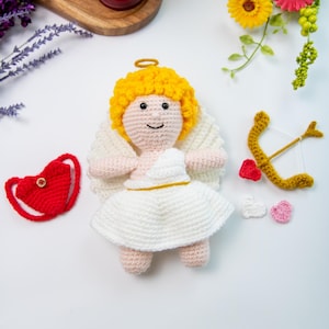 Puede incluir: Un ángel de amigurumi tejido a crochet con cabello amarillo, alas blancas y un vestido blanco. El ángel sostiene un arco y una flecha dorados con un corazón rojo en la flecha. También hay tres corazones pequeños en la superficie blanca.
