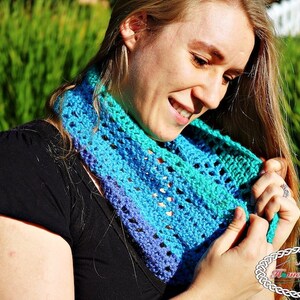 May include: A woman wearing a blue, green and purple crocheted infinity scarf. The scarf is made with a lacy stitch pattern.