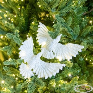 May include: White crochet dove decorations with open wings, set against a green Christmas tree. The handmade doves feature detailed stitching, creating a delicate festive decoration. The tree is illuminated with small, warm white lights.