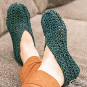 May include: A pair of teal-colored, hand-knitted slippers. The slippers are made with a thick yarn, featuring a textured stitch pattern. The design includes an opening at the top of the foot. The slippers are shown on a person's feet, resting on a gray surface.