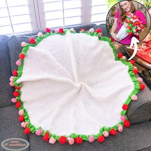 May include: A white crocheted baby blanket with a green border and red and pink rose accents. The blanket is circular and folded on a gray couch. The blanket is handmade.