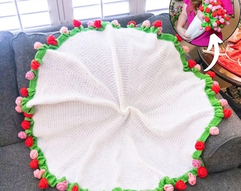 Puede incluir: Una manta de bebé blanca tejida a ganchillo con un borde verde y detalles de rosas rojas y rosas. La manta es circular y está doblada sobre un sofá gris. La manta está hecha a mano.