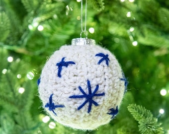 May include: A white crocheted Christmas ornament with blue star and arrow designs. The ornament hangs from a silver metal cap with a silver string. The ornament is hanging in front of a green Christmas tree.