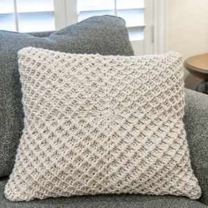 Puede incluir: Una almohada de crochet con un patr&oacute;n de diamantes beige. La almohada est&aacute; sentada en un sof&aacute; gris.
