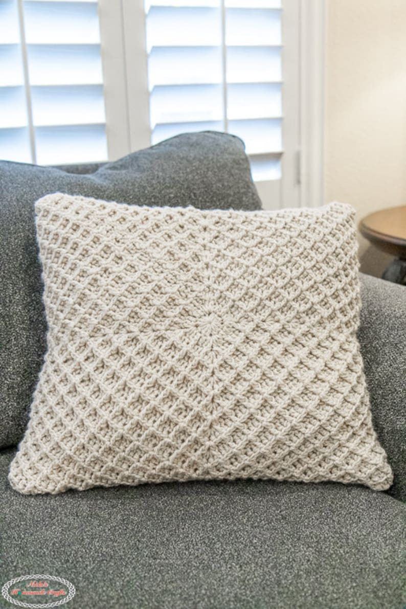 Puede incluir: Una almohada de crochet con un patr&oacute;n de diamantes beige. La almohada est&aacute; sentada en un sof&aacute; gris.