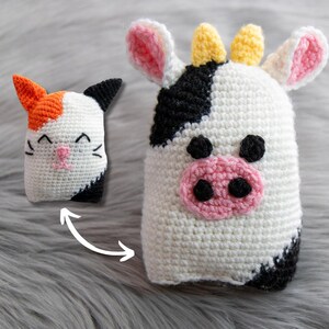 May include: A crocheted cow plushie with black and white spots, pink ears, and a yellow horn. The cow is standing on a gray fuzzy surface. A crocheted cat plushie with orange, white, and black fur is shown in the background.